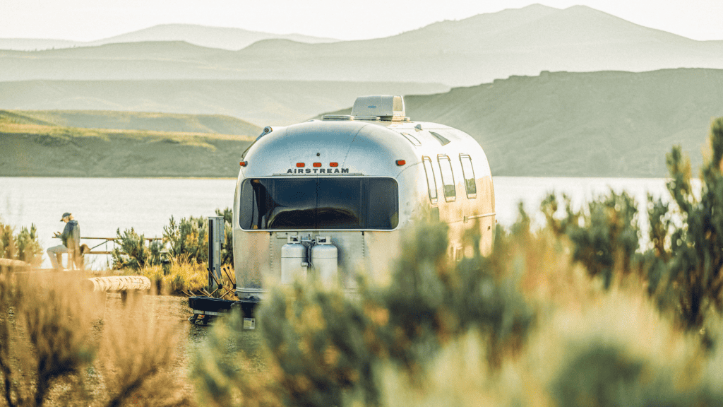 A silver airstream parked next to a lake with mountains in the background.