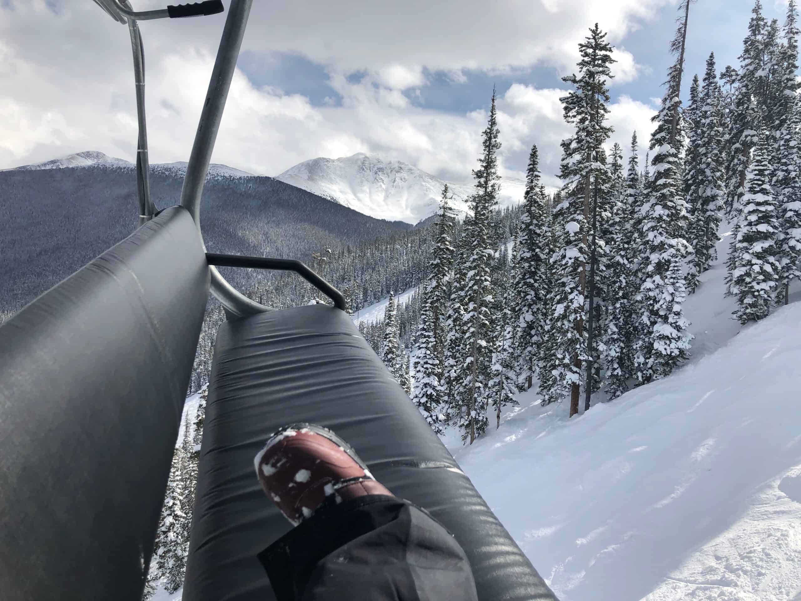 View from a ski lift showing snowy mountains and pine trees, with a person's boot resting on the seat.