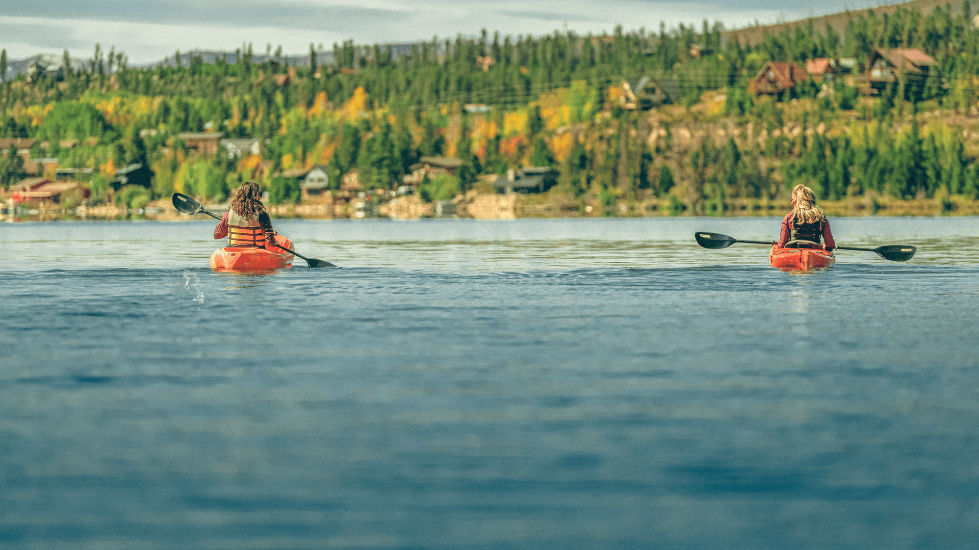 Two people paddling kayaks on a lake with trees in the background.