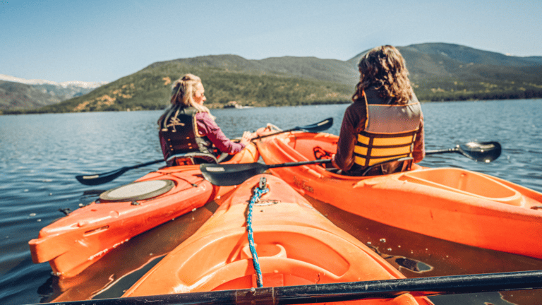 Two women kayaking on a lake with mountains in the background.