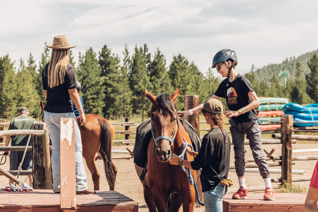 A group of people in a corral learning to horseback ride. One getting on the saddle wears a helmet and the others wear cowboy hats.