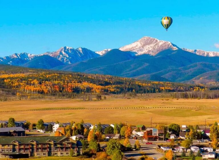 A hot air balloon flies over a town with mountains in the background.