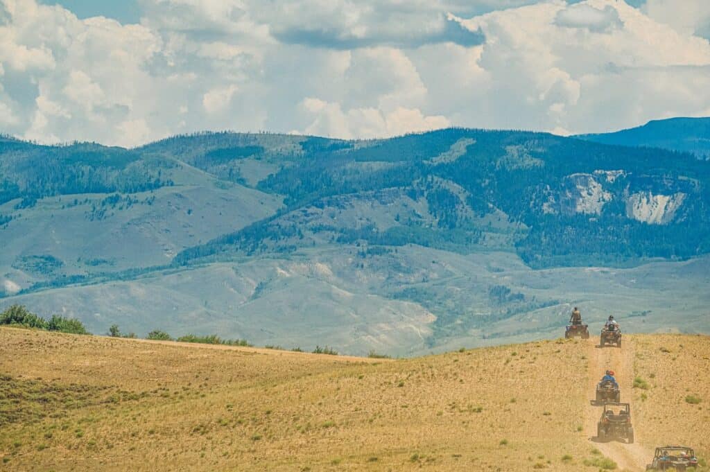 A group of people rides off-road vehicles on a dirt trail in a hilly landscape with mountains in the background under a partly cloudy sky in Kremmling, Colorado.