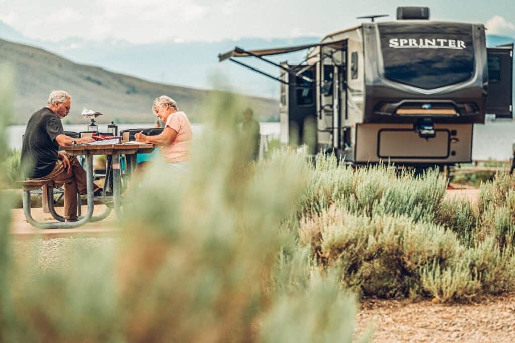 An older couple sits and looks at maps of Grand County at a picnic table at Wolford Mountain Campground. An RV is parked in front of the reservoir in the background.