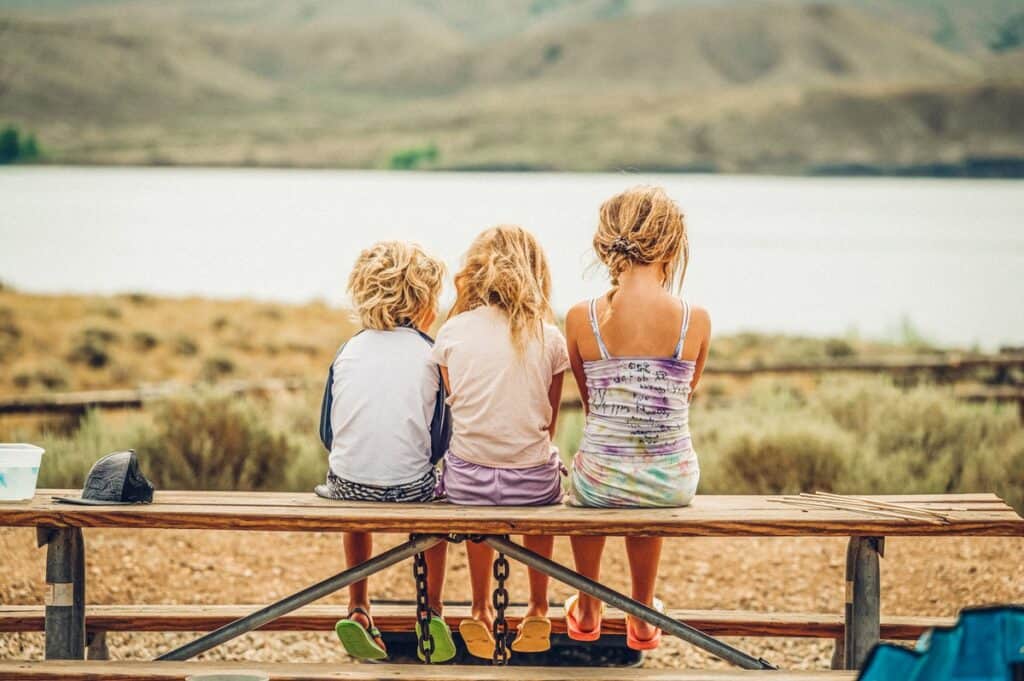 Three children sit on a bench at a Grand County campground and look out on a lake.