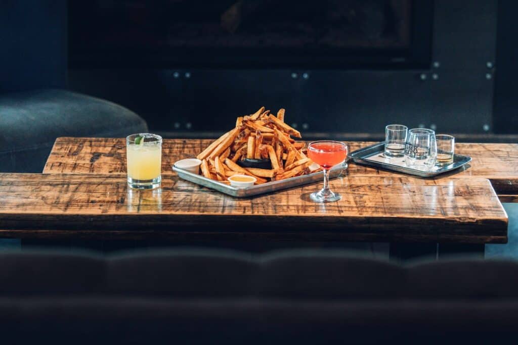 A wooden table with a tray of fries, three dipping sauces, a yellow drink, a red cocktail, and a tray of empty glasses.