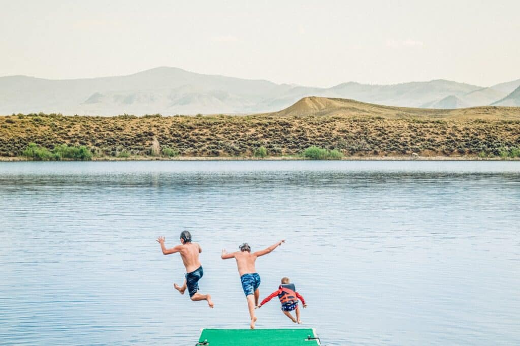 Three kids jump off a dock into a lake in Grand County, with mountains in the background.