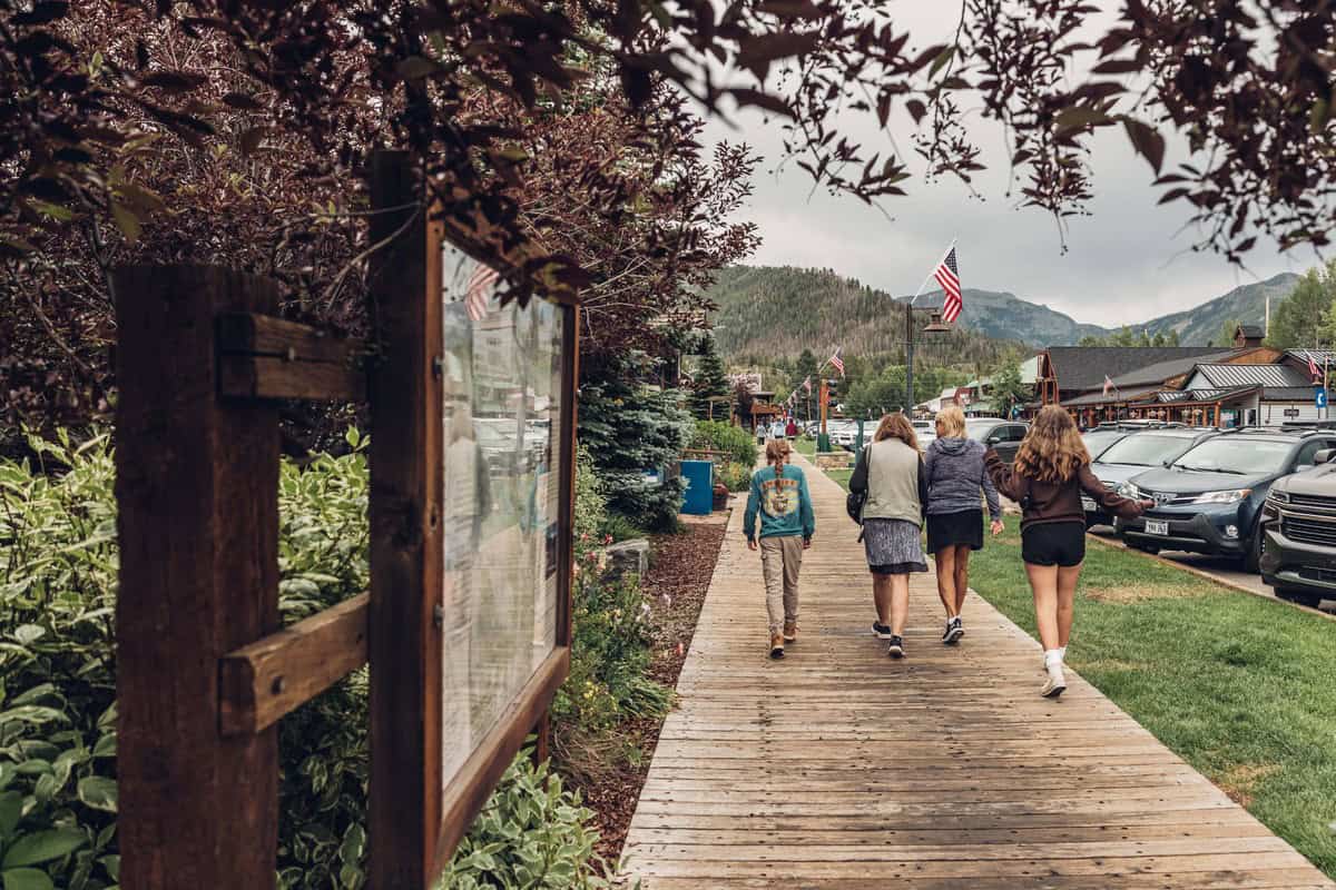 A family walks along a wooden boardwalk in Grand Lake with parked cars on one side and shops in the background. An American flag is visible in the distance.