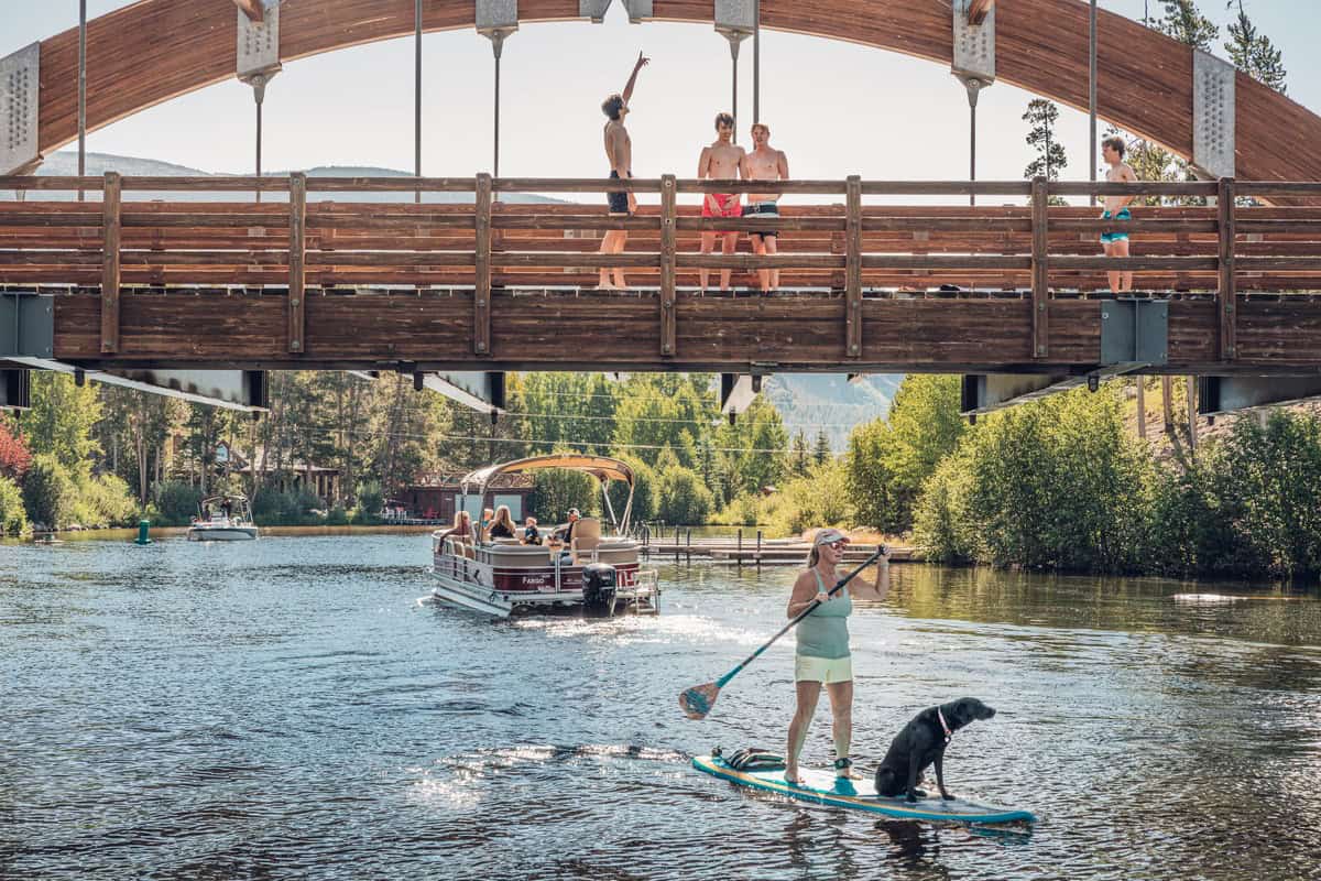 A person paddleboards with a black dog on Grand Lake while people stand on a wooden bridge and a pontoon boat passes underneath.