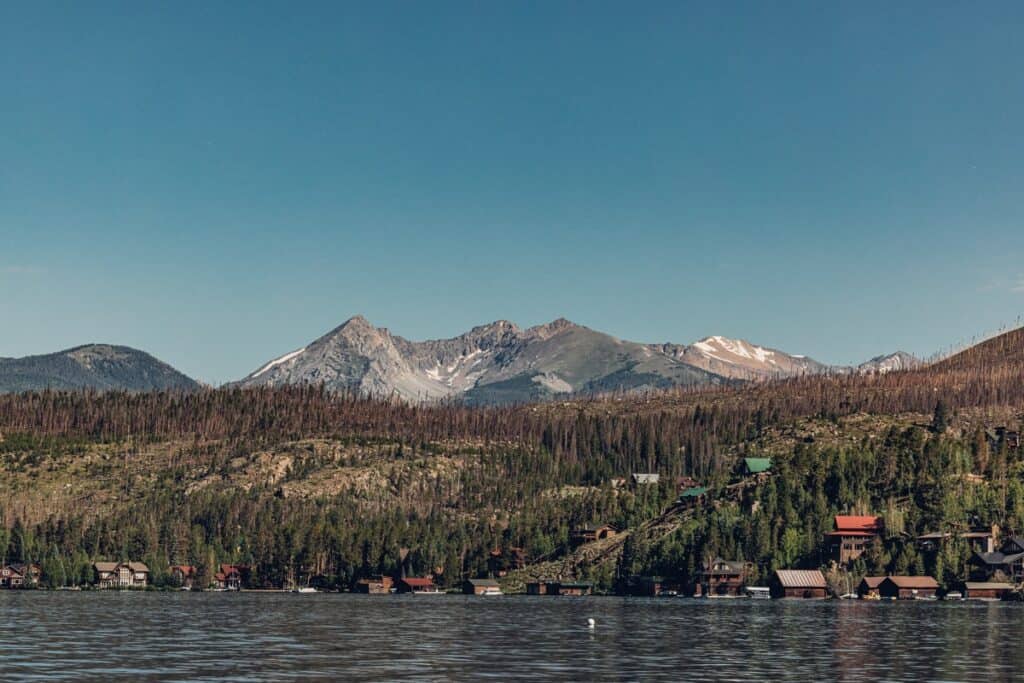 A tranquil Grand Lake is seen in the foreground with a hilly landscape dotted with rustic cabins and a backdrop of snow-capped mountains under a clear blue sky.