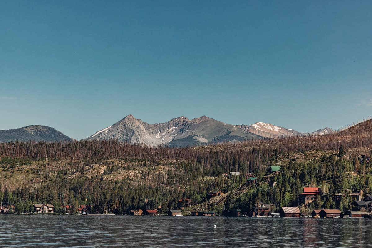 A tranquil Grand Lake is seen in the foreground with a hilly landscape dotted with rustic cabin rentals and a backdrop of snow-capped mountains under a clear blue sky.