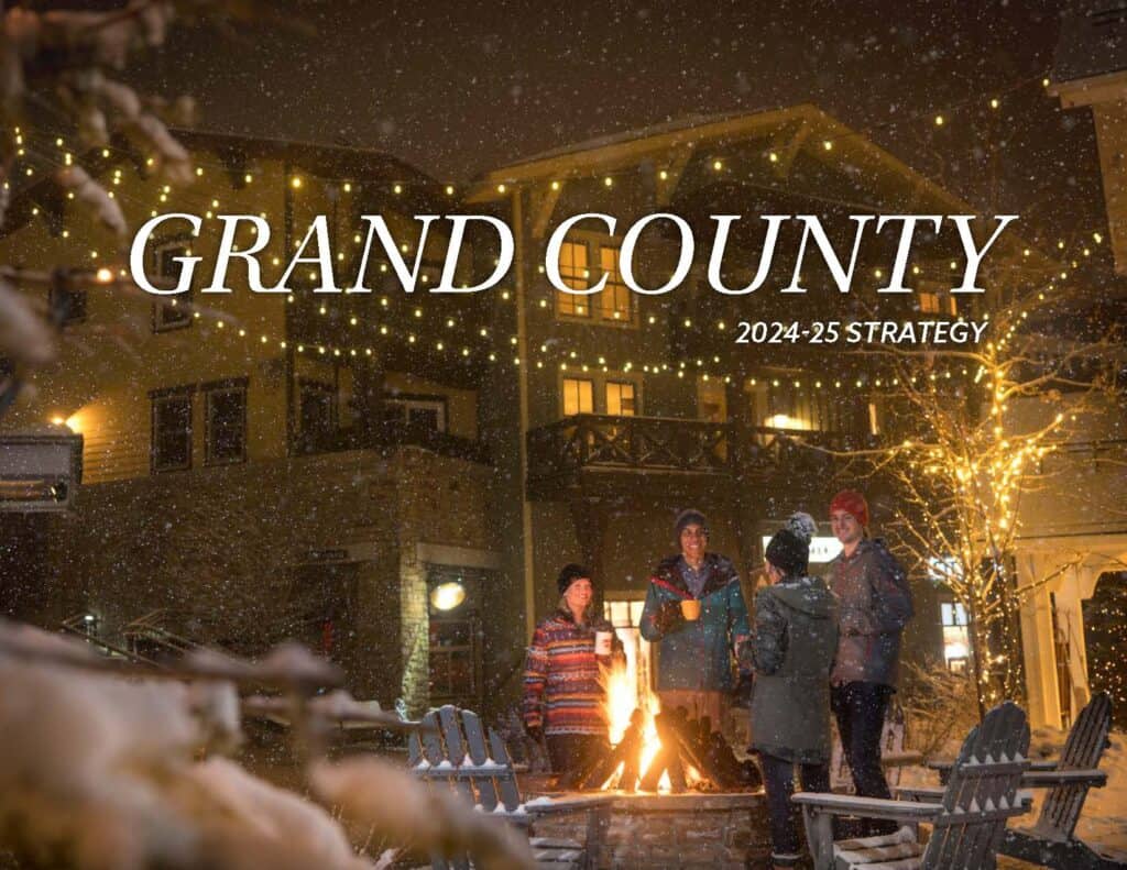 A group of people dressed warmly stand around a fire pit in a snowy, festively lit outdoor area, with "Grand County 2024-25 Strategy" text overlay.