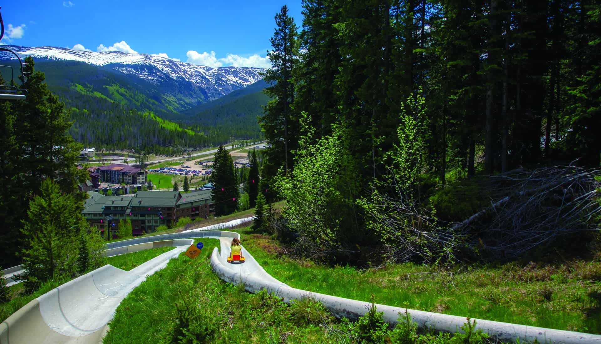 A person rides down an outdoor alpine slide near Granby, CO amid green trees, with a mountainous landscape and buildings in the background.