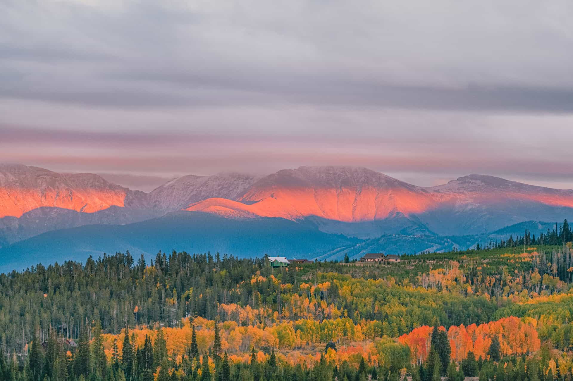 Scenic view of a Rocky Mountain range at sunrise, with peaks illuminated in orange hues. In the foreground, a forest with autumn-colored trees stretches across the landscape.