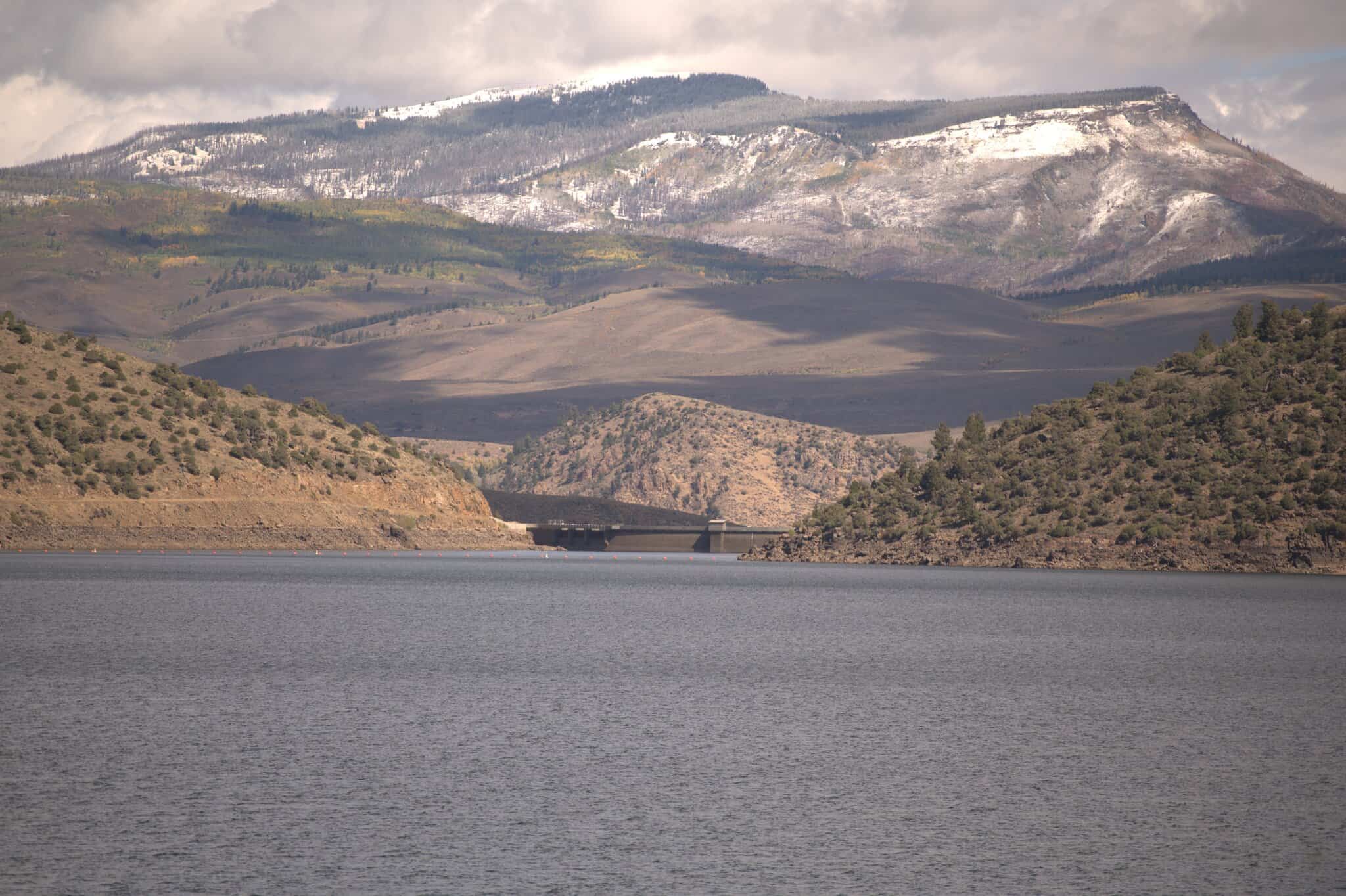 Mountain landscape with Williams Fork Reservoir in the foreground, marked by rugged hills and a concrete bridge in the distance below snow-flecked mountains. Cloudy sky overhead.