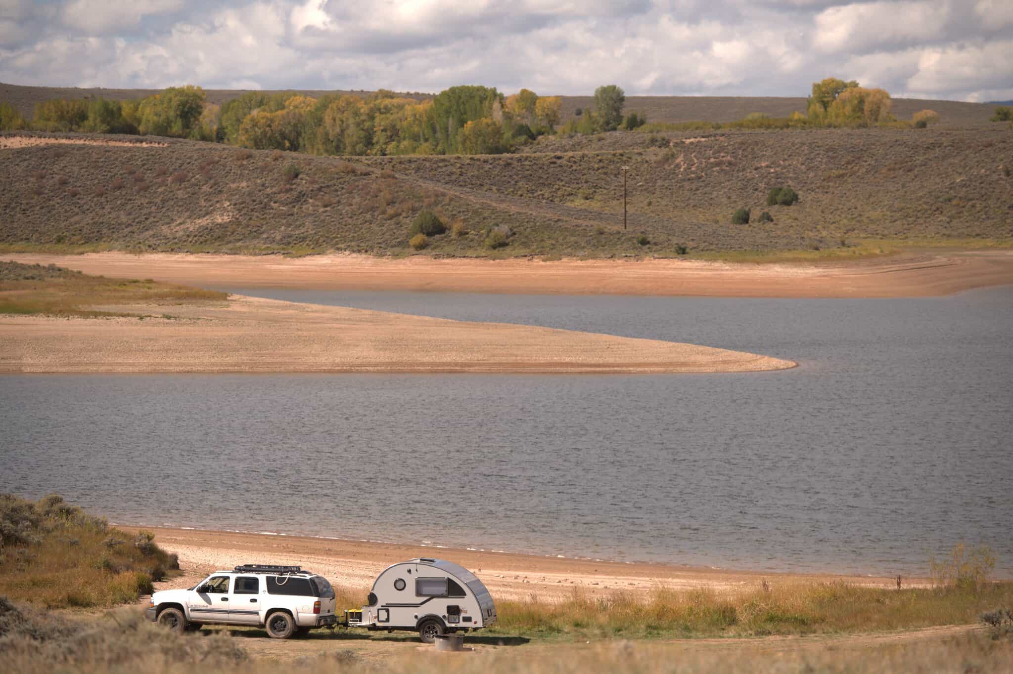 A white truck with a small camper trailer is parked near a calm Williams Fork Reservoir, surrounded by hills and sparse foliage under a cloudy sky.