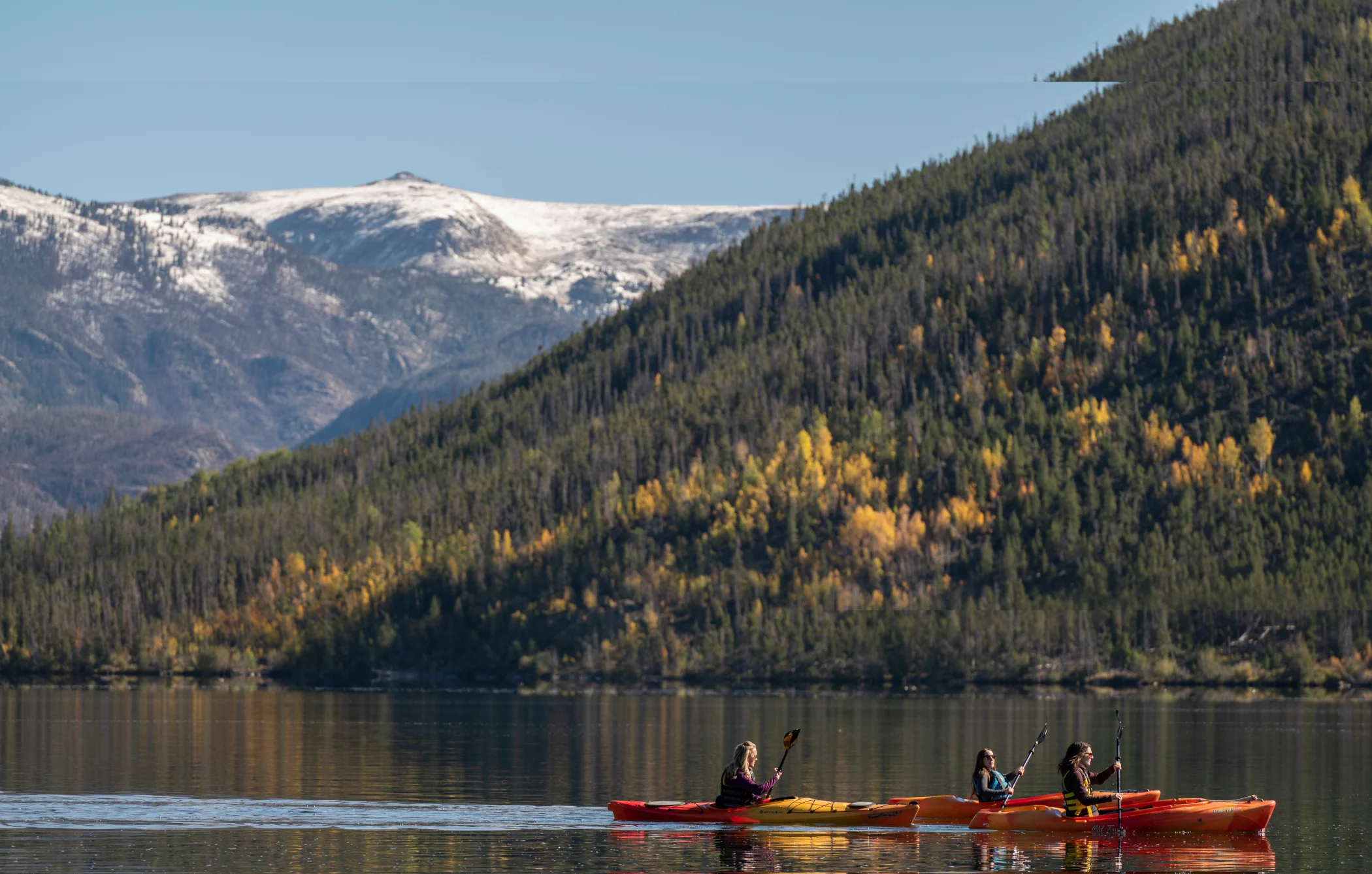 Three people kayak on a calm lake near forested mountains with some snow-capped peaks in the background under a clear sky.