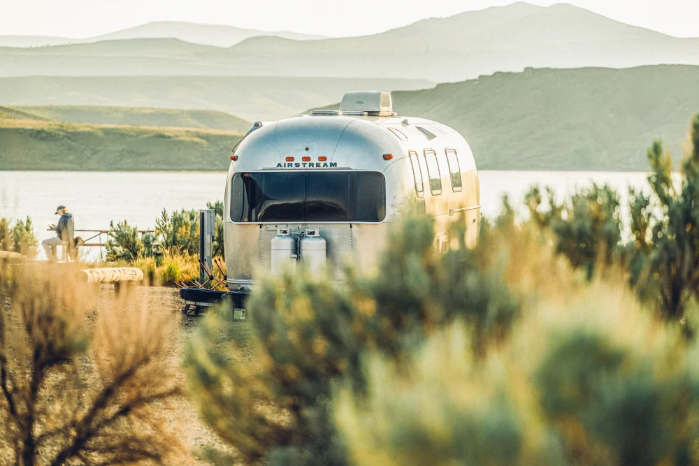 A silver Airstream trailer is parked near a lake with mountains in the background. A person walks nearby, partially obscured by vegetation.