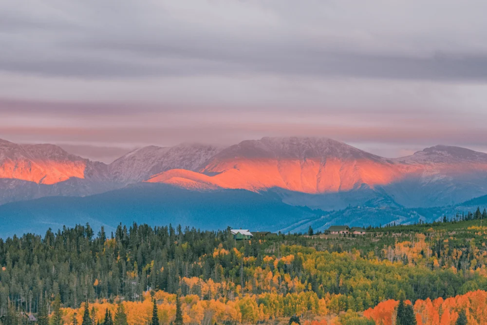 A scenic view of a mountain range with autumn foliage in the foreground and the mountains illuminated by the pink and orange hues of a setting or rising sun.