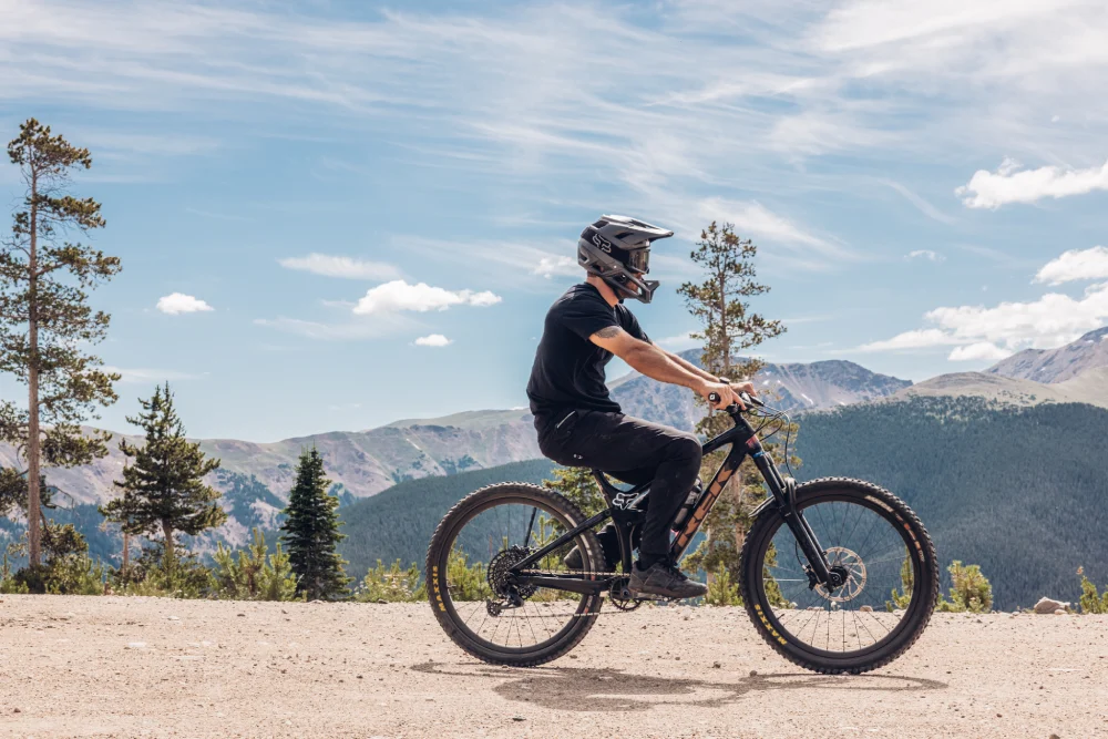 A person wearing a helmet and black clothing rides a mountain bike on a dirt path with a scenic backdrop of trees and mountains under a partly cloudy sky.