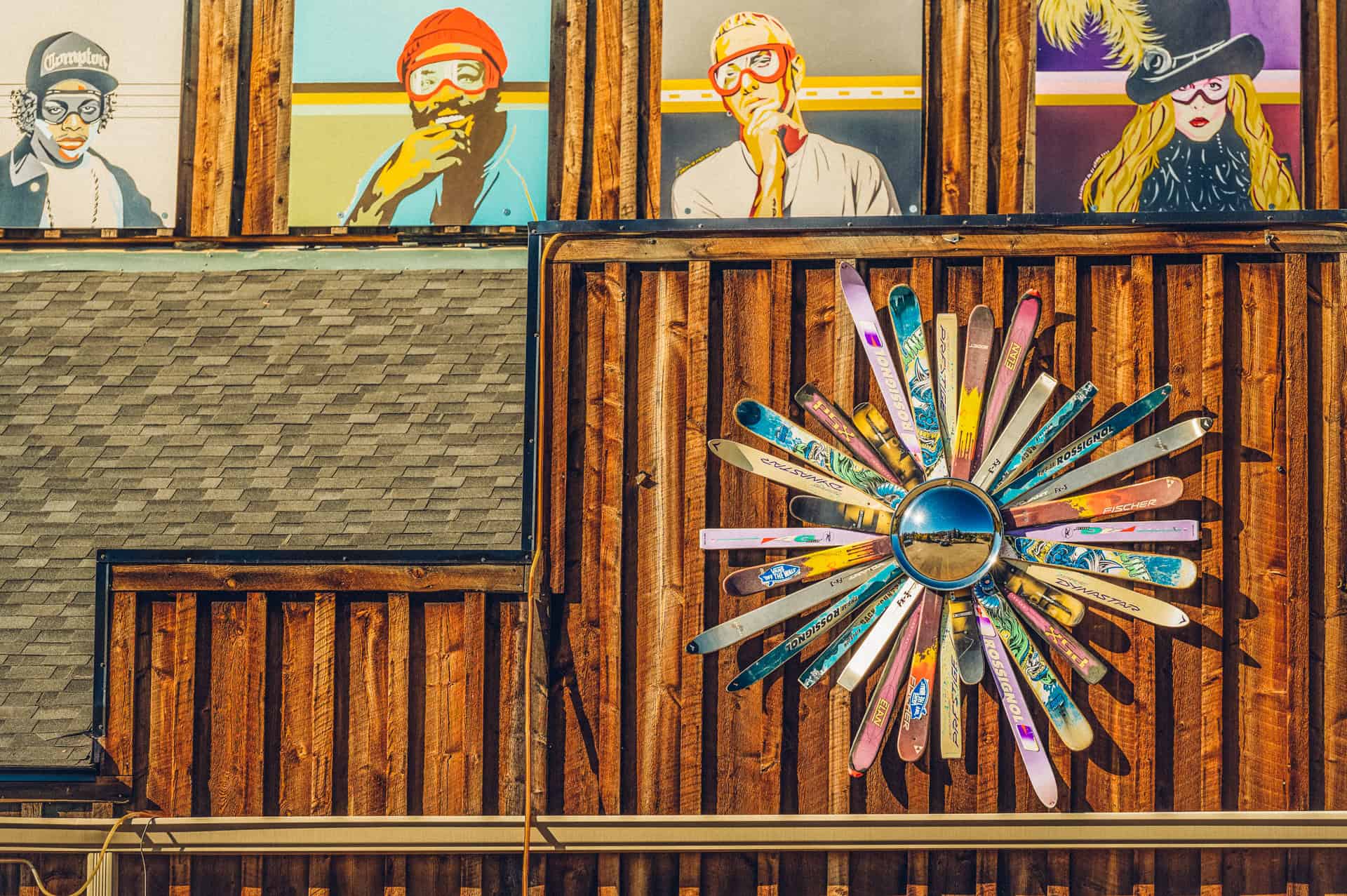 A wooden building in Cooper Creek Square features colorful portraits of people above a circular art piece made from skis.