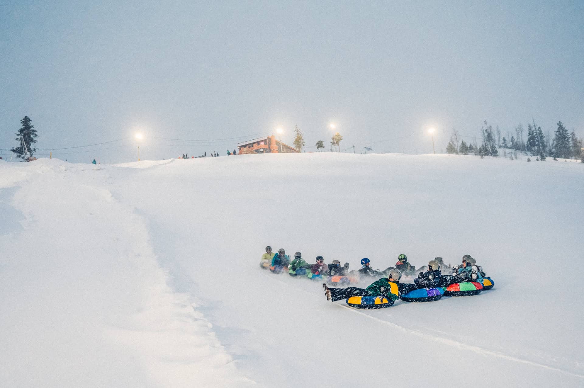 Group of people tubing down a snowy hill near Winter Park, with a building and lights in the background.