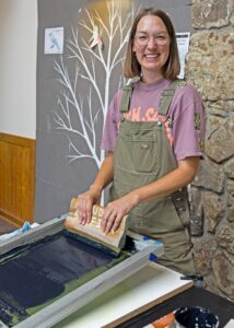 A person in overalls and a t-shirt smiles while working on a screen printing project indoors.