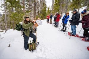 A park ranger in snowshoes shows a wildlife guide to a group of people standing on a snowy trail in a forest.